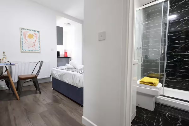 Modern bedroom with desk, chair, bed visible through doorway, and en-suite bathroom with black tile shower