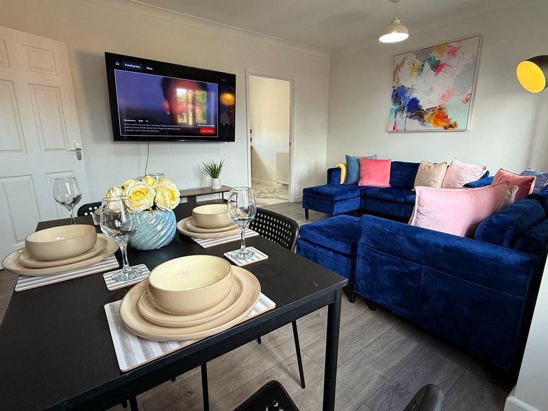 Open-plan living and dining area with blue velvet sofa, dining table set for four, wall-mounted TV, and abstract wall art