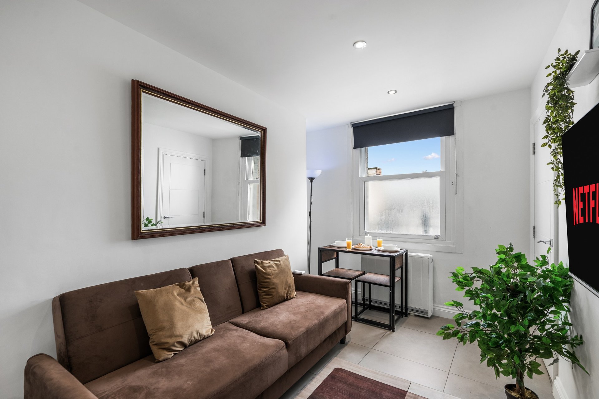 Bright living room with brown sofa, wall mirror, small desk, potted plants and window