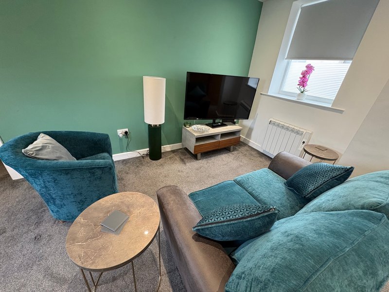 Modern living room with teal upholstered seating, wooden coffee table, TV, and green accent wall