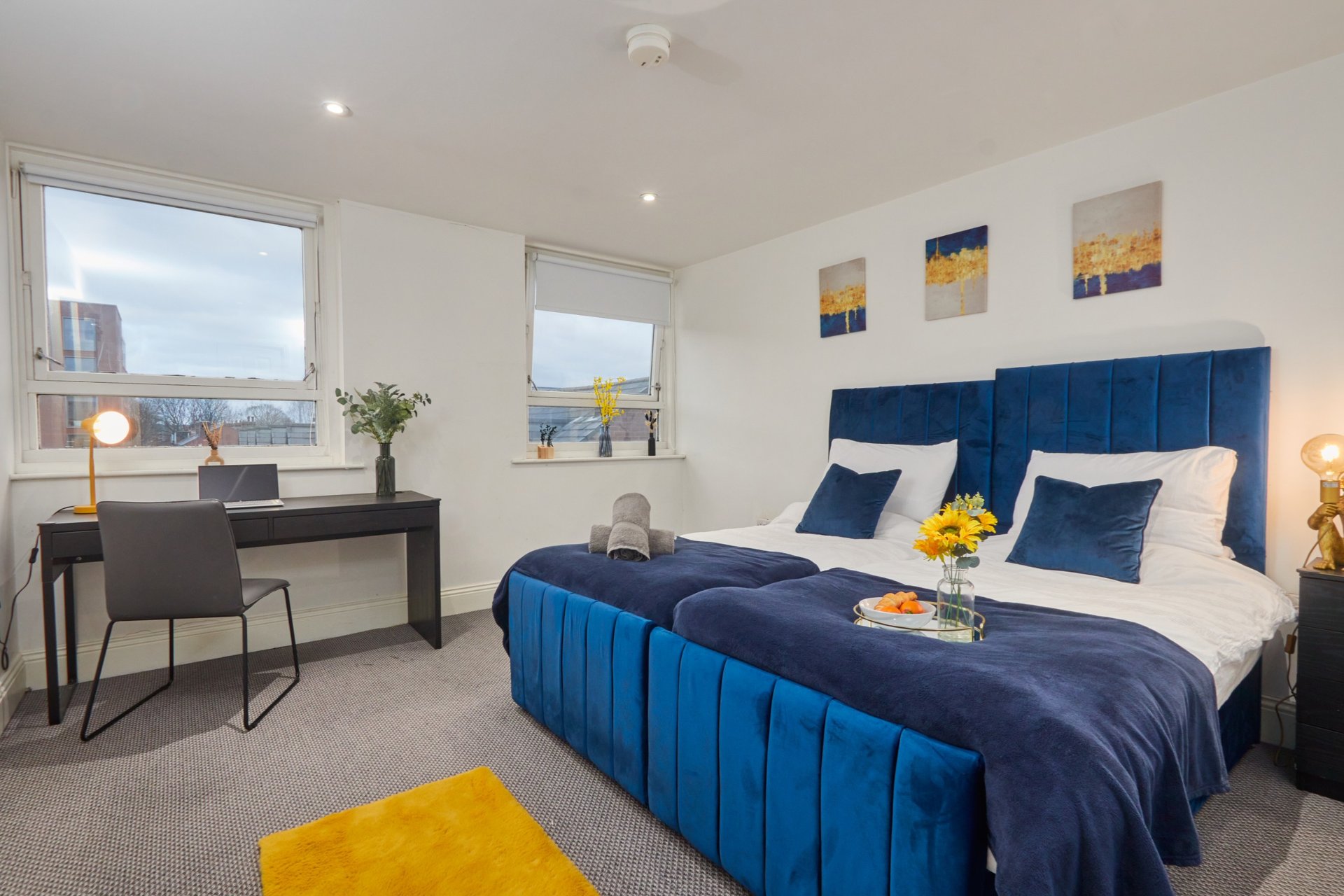 Modern bedroom with blue upholstered bed, desk area, windows, and yellow accent furniture