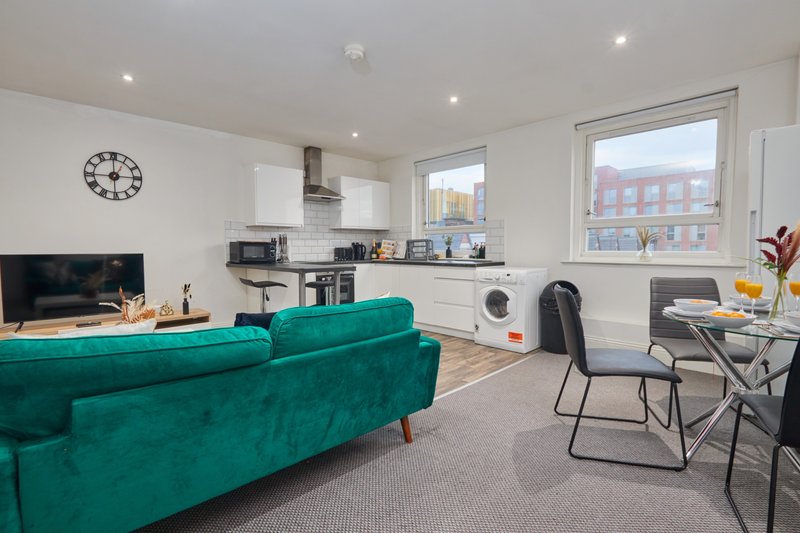 Open-plan living area with teal sofa, dining table, kitchen, and wall-mounted clock