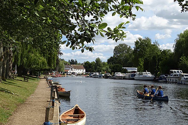 Riverside towpath with moored boats, tree-lined banks, and waterfront buildings in distance