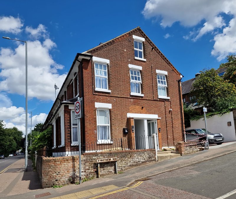 Red brick building exterior with white-framed windows, street frontage and parked vehicles visible