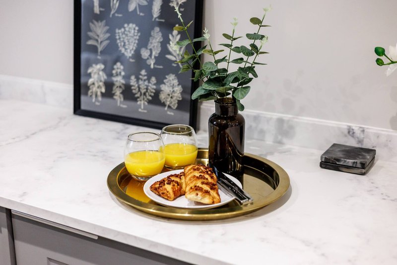 Kitchen counter with marble top, brass tray holding glasses of juice, pastries, vase with greenery and botanical wall art