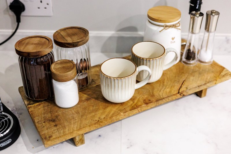 Kitchen counter with wooden tray holding ceramic mugs, glass jars with wooden lids, and toiletries