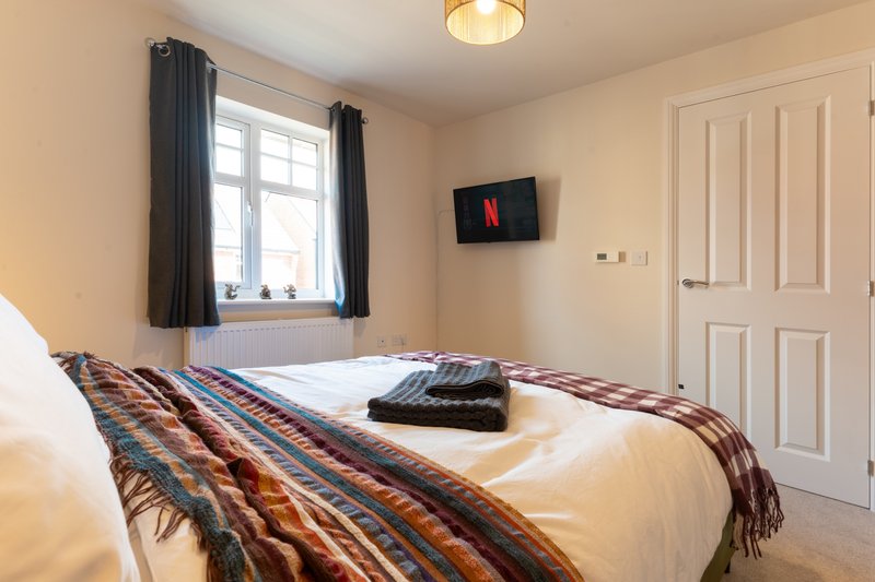 Bright bedroom with double bed, patterned throw blanket, window with dark curtains, wall-mounted TV, and cream-colored walls