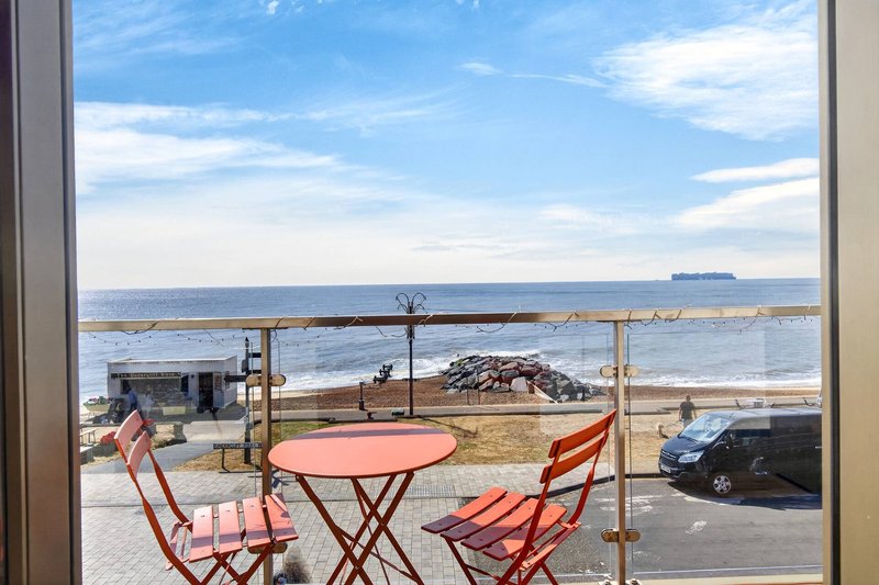 Beachfront view from balcony with red chairs, small table, and seaside promenade beyond