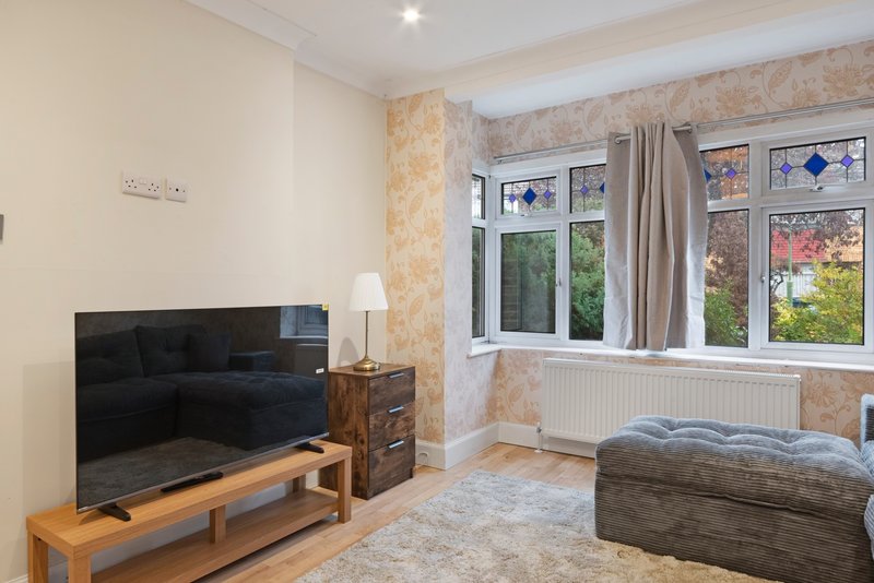 Living room with flat-screen TV, wooden TV stand, upholstered armchair, and large windows with garden views