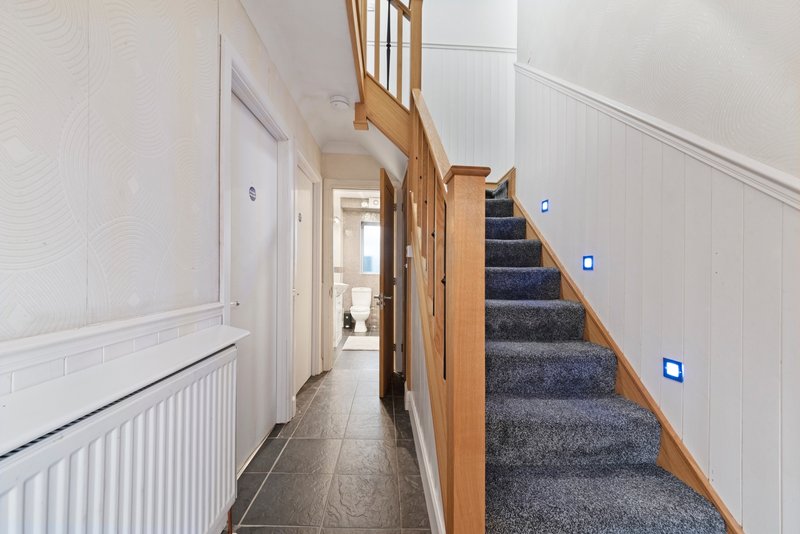 Hallway with staircase, wooden banister, white walls, dark flooring and blue lighting.