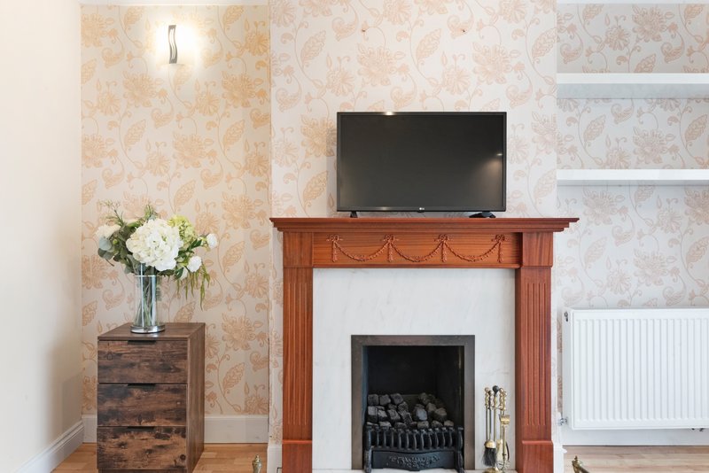 Living room with fireplace, TV on mantelpiece, floral wallpaper and white flowers in vase