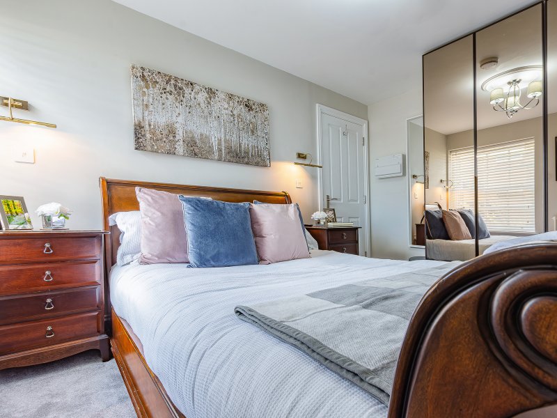 Bedroom with wooden bed frame, colorful pillows, bedside table, dresser, and mirrored wardrobe
