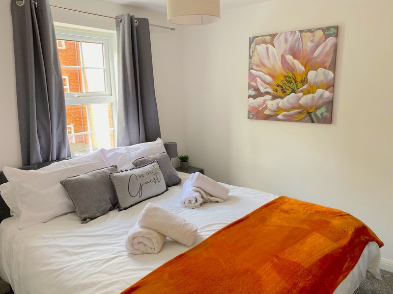 Bright bedroom with double bed, white bedding, orange throw, gray curtains and floral wall art