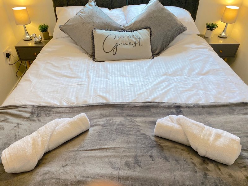 Modern bedroom with white bed, decorative pillows, bedside lamps, and rolled towels