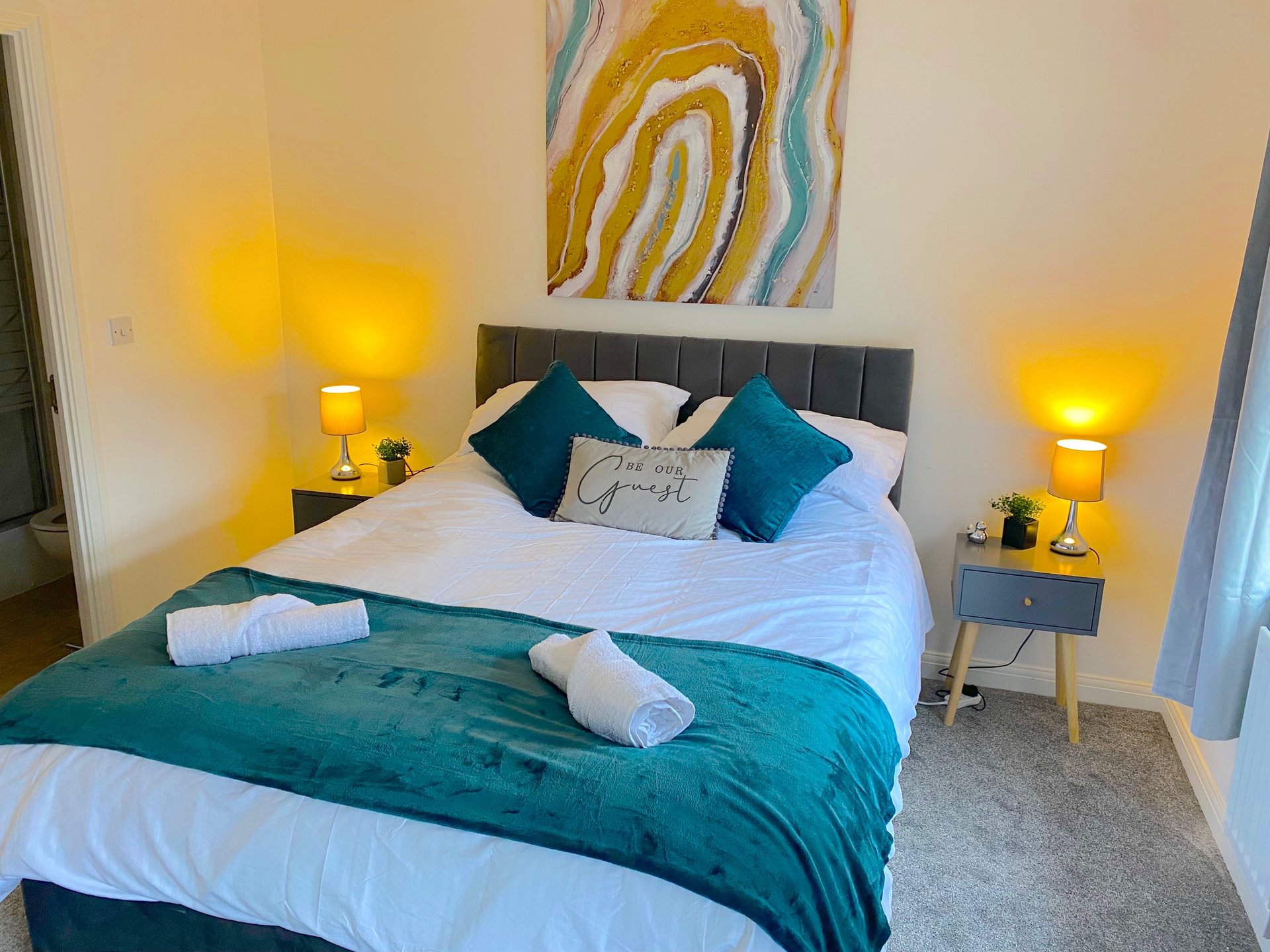 Modern bedroom with teal and white bedding, yellow lamps, and abstract wall art