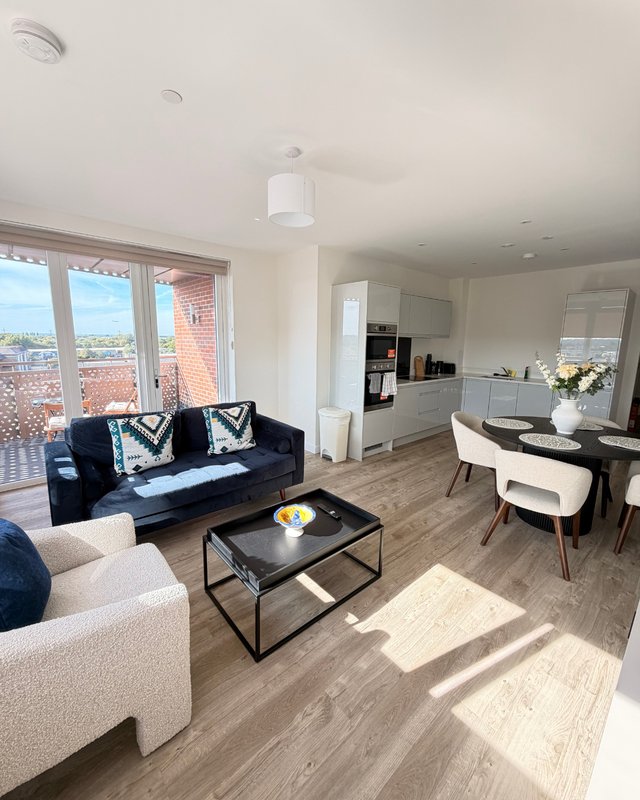 Open-plan living area with black sofa, cream armchair, dining table and kitchen, large windows with city views