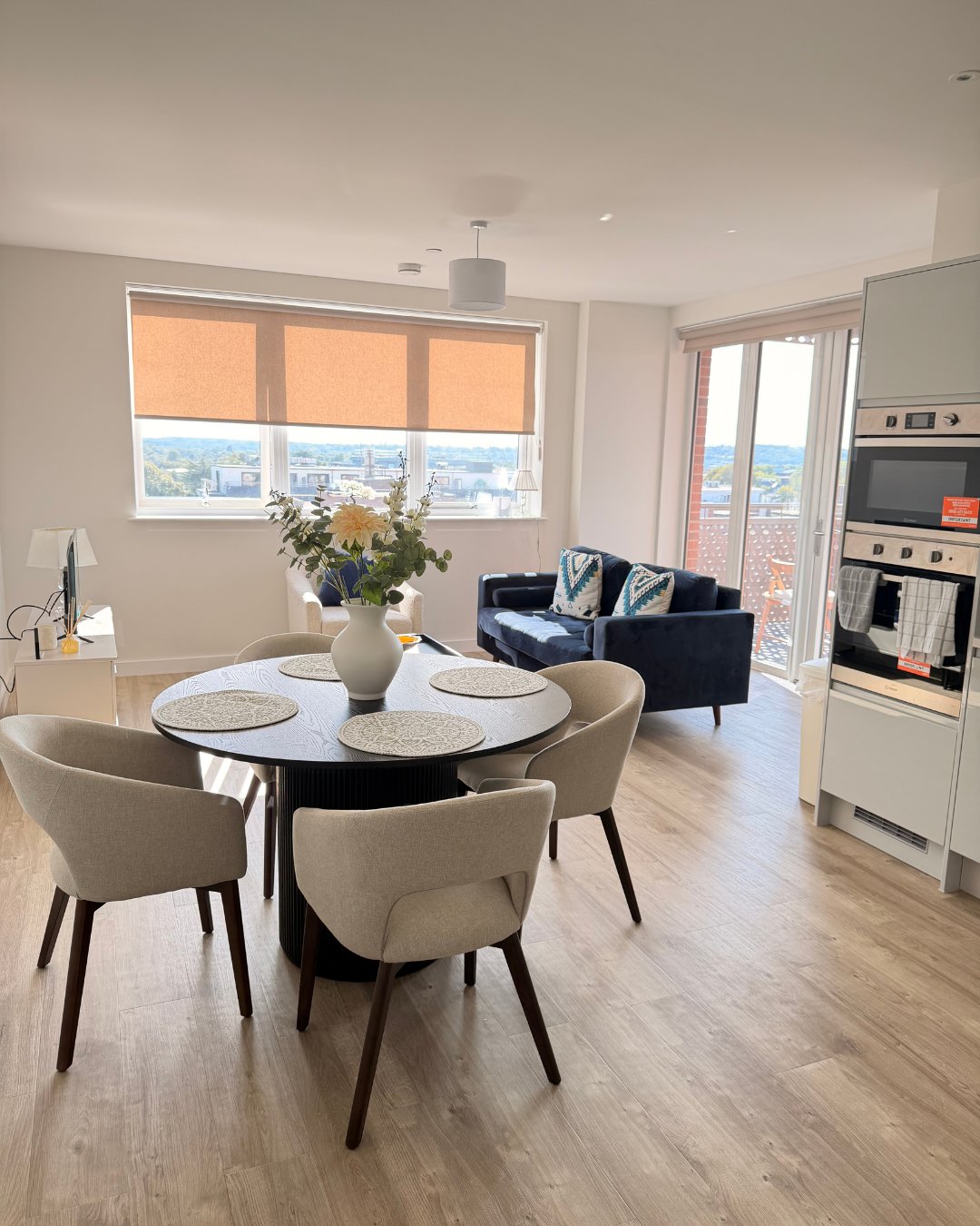 Open-plan kitchen and dining area with round table, cream chairs, dark sofa, and city views through large windows