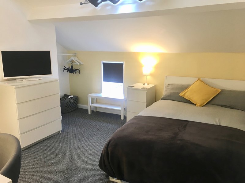 Bedroom with double bed, white dresser, TV, yellow accent wall and bedside lamp