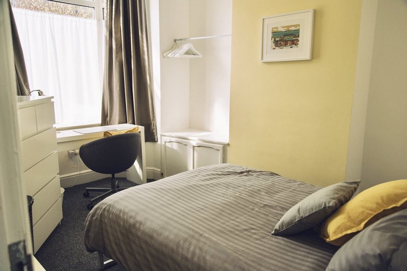 Bedroom with double bed, yellow walls, desk chair, window with curtains and framed artwork