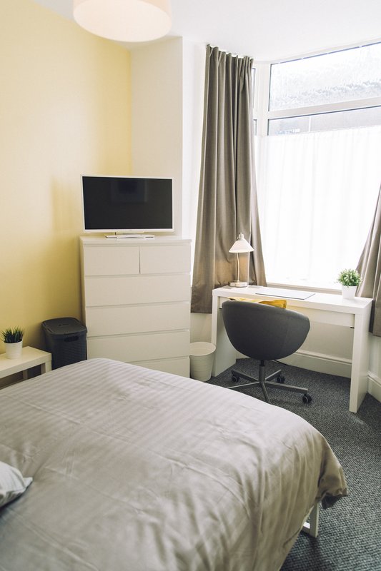 Bedroom with double bed, TV on white chest of drawers, desk chair, and window with grey curtains