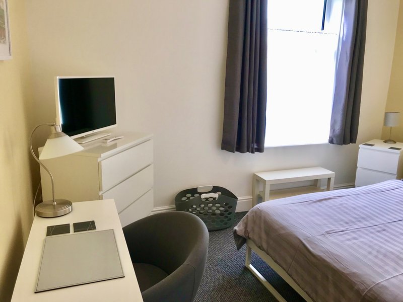 Compact bedroom with single bed, desk, TV, chair and window with curtains