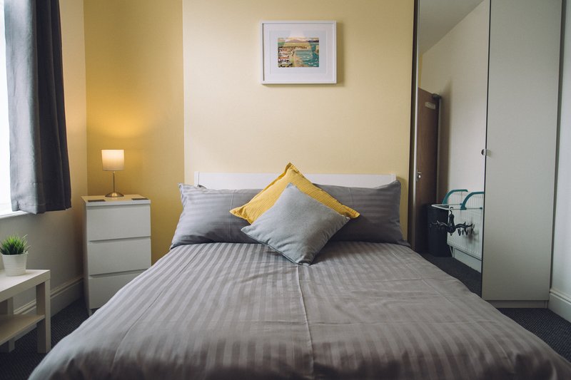 Bedroom with double bed, gray bedding, yellow and gray pillows, bedside lamp and white nightstand