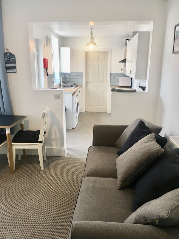 Open-plan living area with sofa, chair, kitchen, and doorway to bedroom beyond