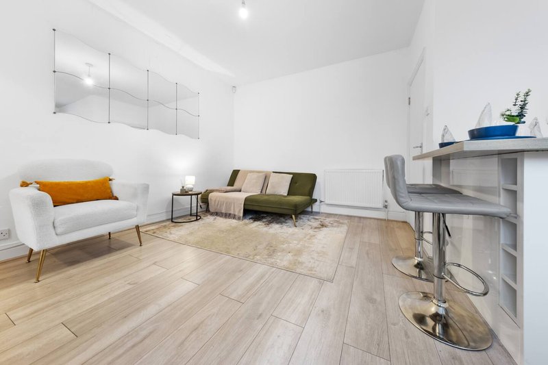 Bright modern living room with white walls, wooden flooring, white armchair, olive sofa, and kitchen counter