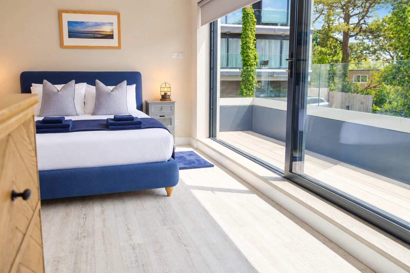 Bright modern bedroom with navy blue bed, white bedding, wooden flooring and large sliding glass doors