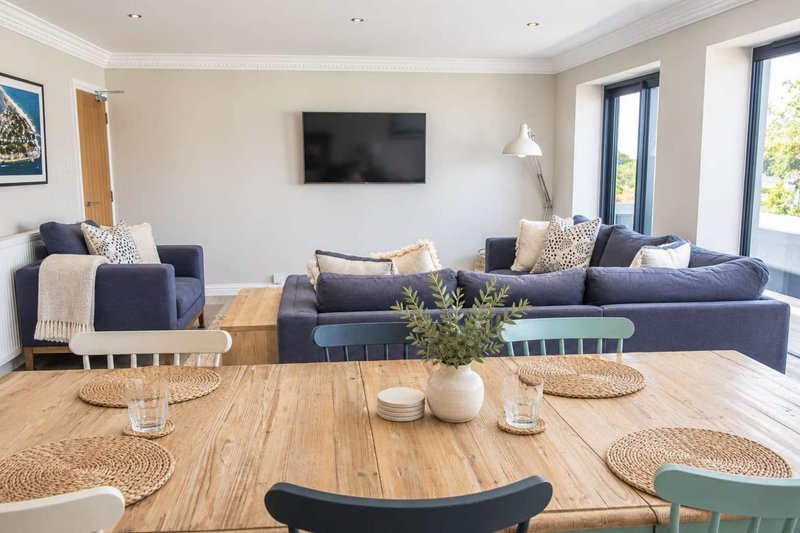 Open-plan living and dining area with wooden table, blue sofas, wall-mounted TV and large windows