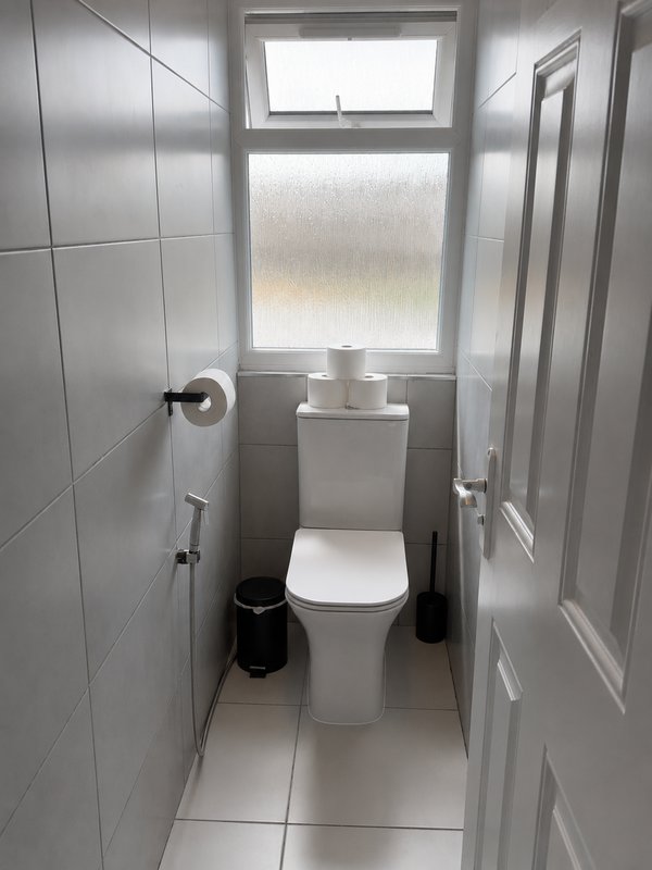 Small bathroom with white toilet, tiled walls, window and black waste bin