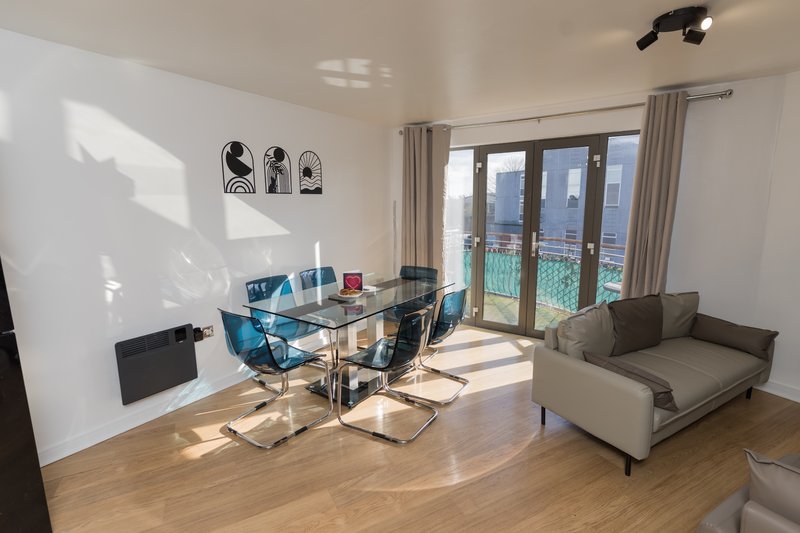 Modern open-plan living and workspace with glass desk, blue chairs, sofa, and balcony with city views
