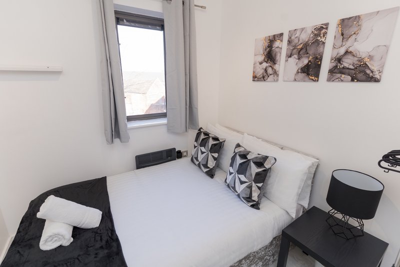 Bright modern bedroom with white double bed, patterned cushions, window with grey curtains, wall art