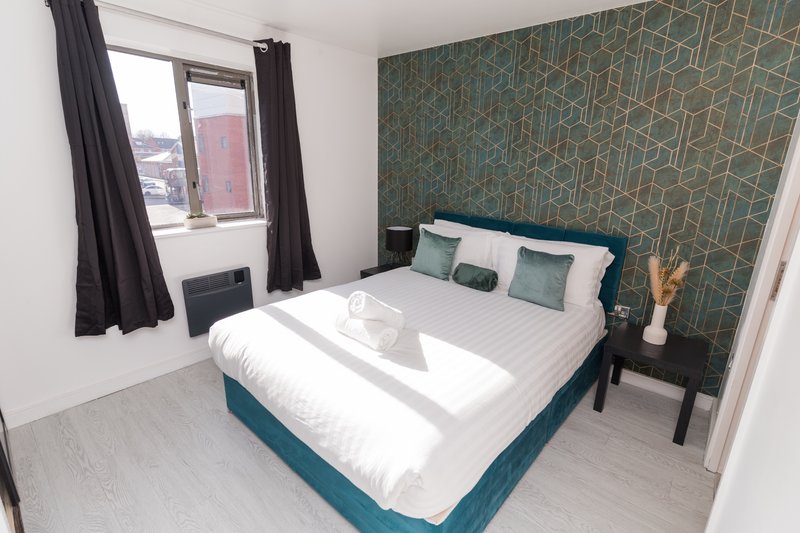 Modern bedroom with teal upholstered bed, geometric wallpaper, window with black curtains and city view