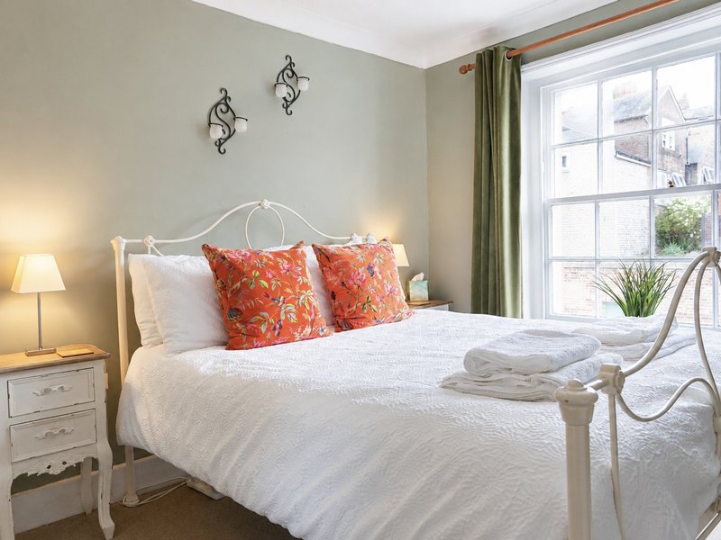 Bright bedroom with white metal bed frame, orange patterned pillows, white bedding, and large window with garden view