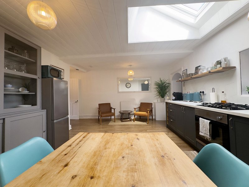 Modern open-plan kitchen and living area with wooden dining table, turquoise chairs, skylights, and contemporary furnishings
