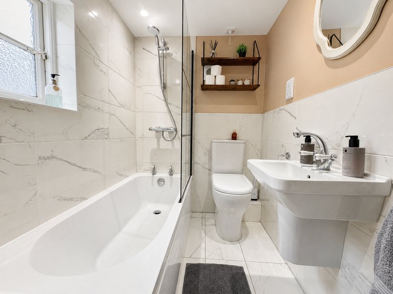 Compact bathroom with bath, shower screen, toilet, sink and wall-mounted shelving.