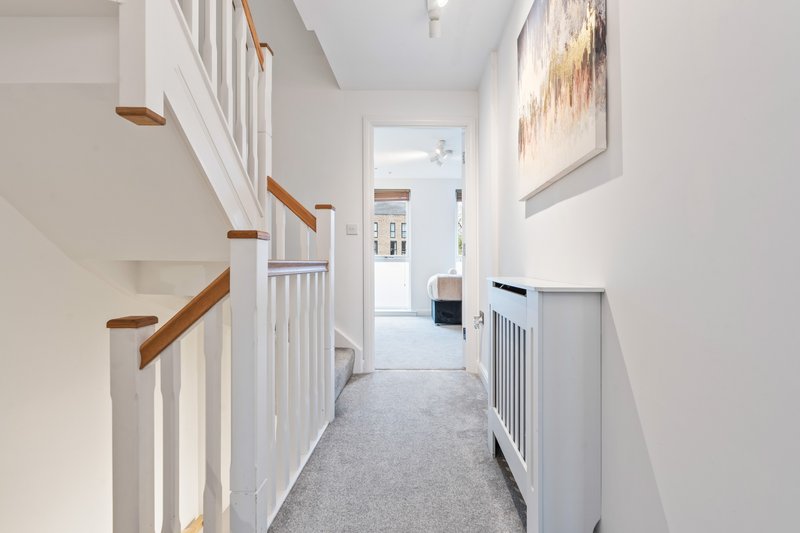 Bright hallway with white walls, wooden stair railing, grey carpet, and framed artwork