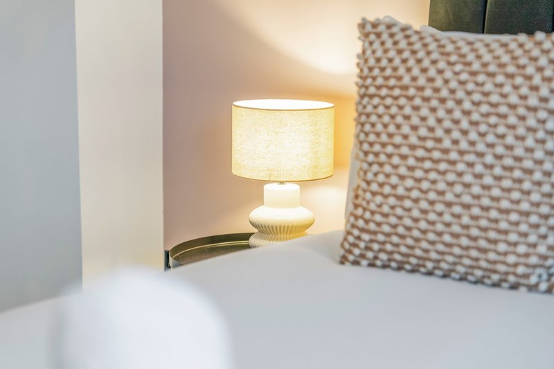 Bedside table lamp with cream-colored cylindrical shade and decorative patterned cushion