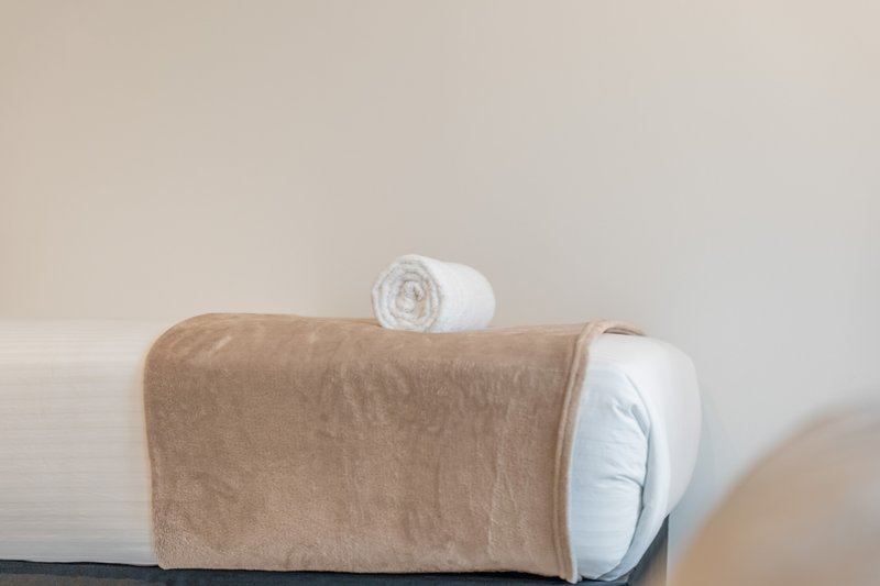 Bed with white pillows, tan throw blanket, and rolled towels arranged on top