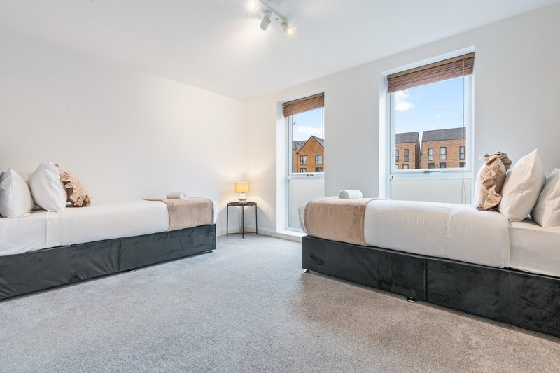 Twin bedroom with two beds, cushions, side table, lamps, and large windows overlooking street