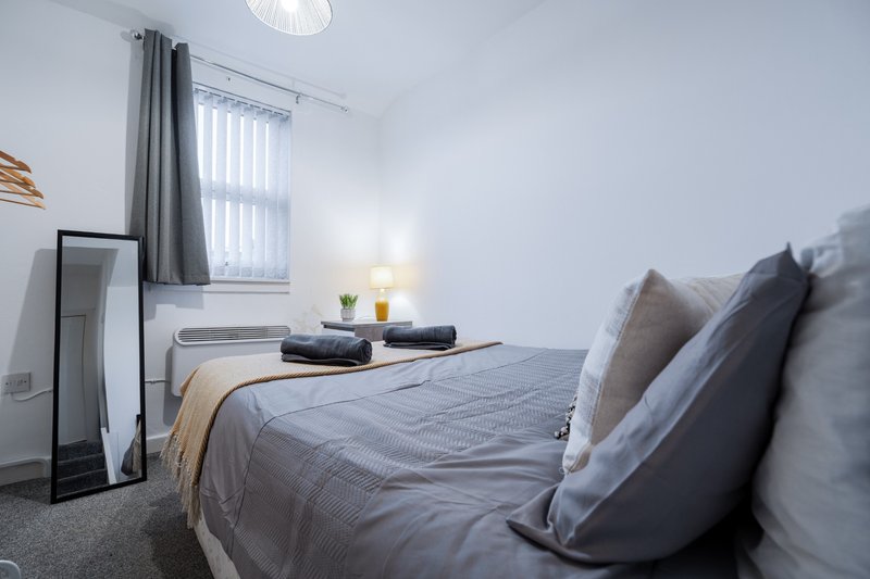 Bedroom with double bed, gray bedding, pillows, bedside lamp and window with curtains