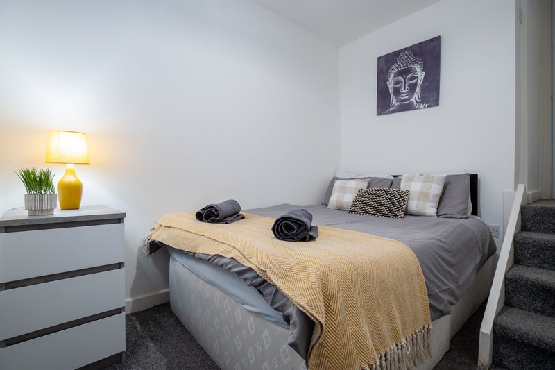Bedroom with double bed, yellow throw, headboard pillows, bedside lamp and wall art