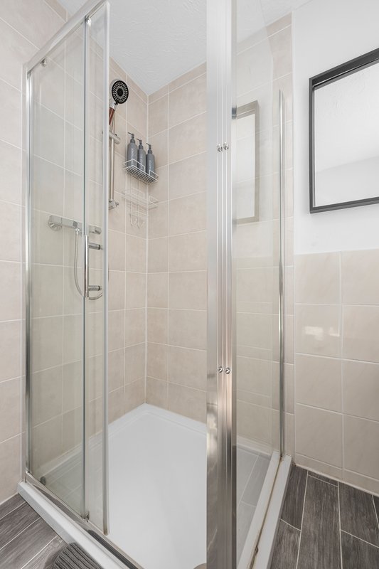 Modern shower enclosure with glass doors, beige tile walls, and dark wood-look flooring.
