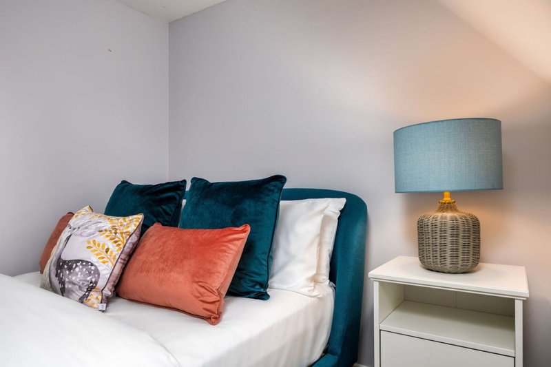 Bedroom with teal upholstered bed, colorful cushions, white bedding, and bedside table with lamp