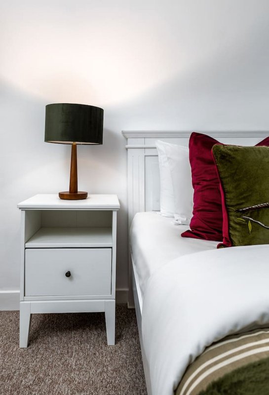 Bedroom with white nightstand, table lamp, and bed with burgundy and olive cushions