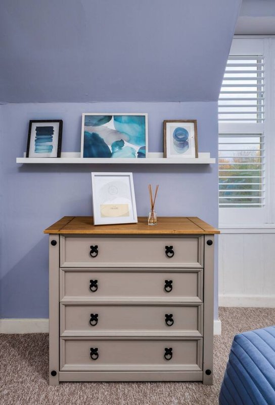 Bedroom with pale purple walls, wooden chest of drawers, floating shelf with framed artwork, and window with shutters