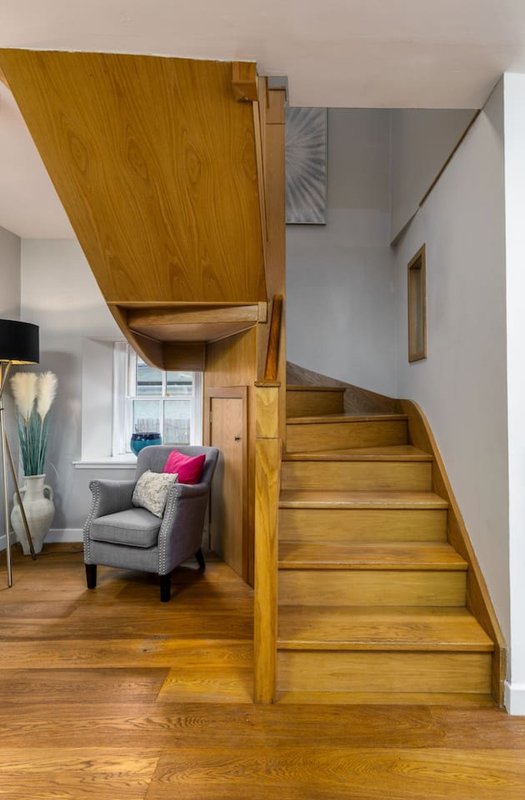 Modern living area with wooden staircase, gray armchair with pink cushion, and open-plan layout
