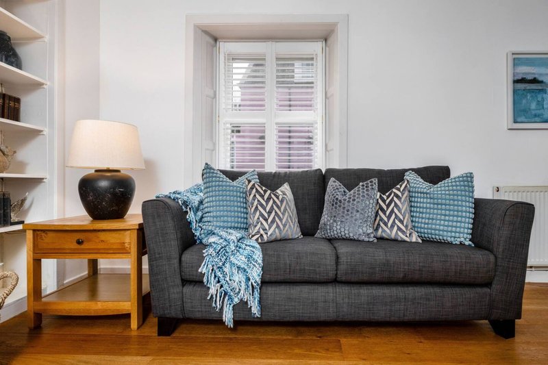 Living room with dark grey sofa, patterned cushions, wooden side table with lamp, and shuttered window