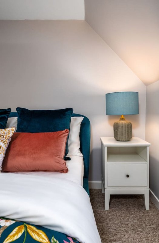 Bedroom with blue-upholstered bed, white bedside table, lamp, and colorful cushions